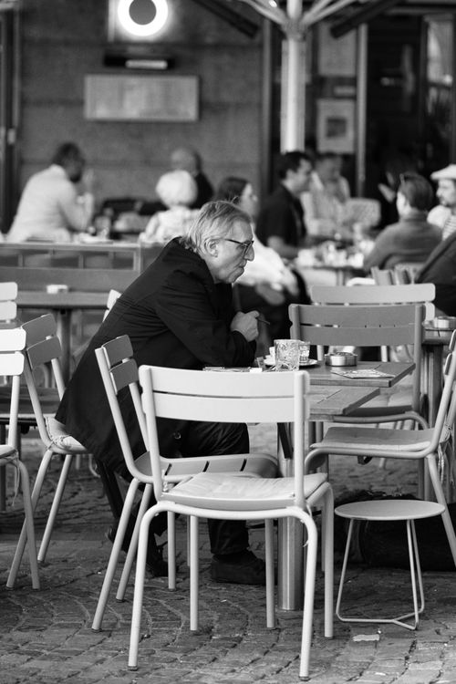 A man in a suit sits alone at an outdoor café table, surrounded by empty chairs, with people in the background.